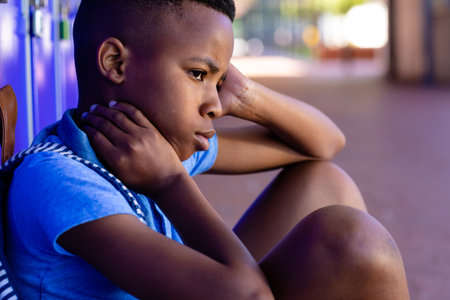 Close up of sad african american schoolboy sitting by lockers in school, with copy space. Education, childhood, elementary school, inclusivity and learning concept.の写真素材