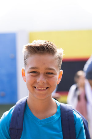 Vertical portrait of smiling caucasian elementary schoolboy in school playground, copy space. Education, inclusivity, elementary school and learning concept.の写真素材