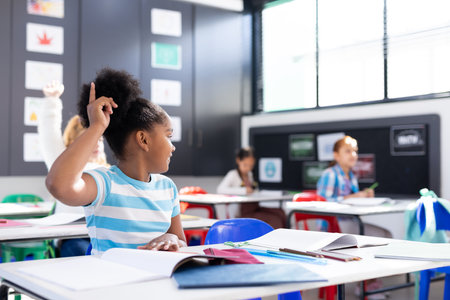 African american schoolgirl sitting at desk raising hand in elementary school classroom, copy space. Education, inclusivity, elementary school and learning concept.の写真素材