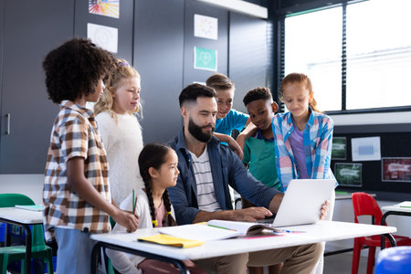 Diverse elementary schoolchildren and male teacher using laptop at desk in classroom, copy space....