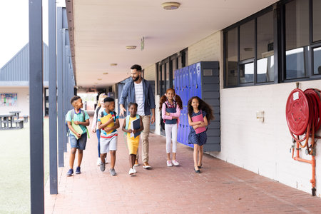 Diverse, happy male teacher and elementary schoolchildren walking in school corridor, copy space. Education, inclusivity, elementary school and learning concept.の写真素材