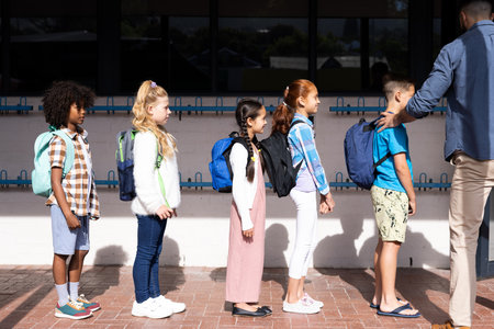 Diverse male teacher and elementary schoolchildren with schoolbags queuing in school playground. Education, inclusivity, elementary school and learning concept.の写真素材