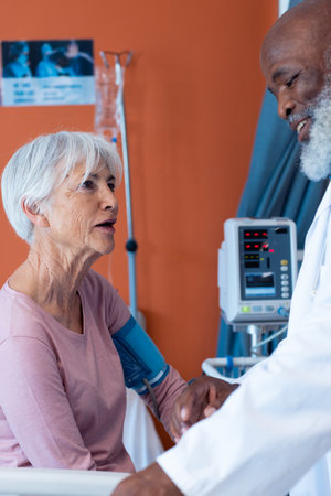 Vertical of happy diverse senior male doctor taking blood pressure of female patient, copy space. Hospital, medical and healthcare services.の写真素材