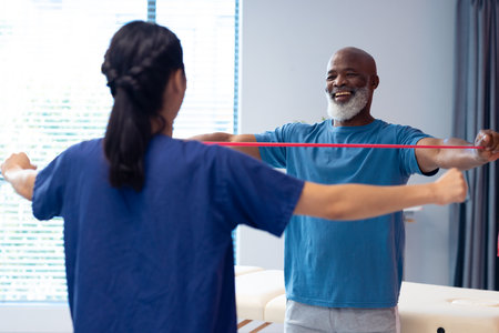 Diverse female physiotherapist helping smiling senior male patient exercise with band, copy space....
