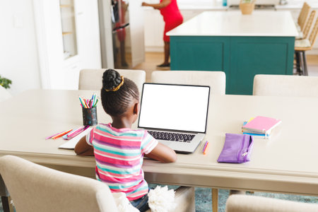 Rear view of african american girl with hair bun studying online over laptop on table at home. Copy space, unaltered, education, e-learning, childhood, book, pencil and wireless technology concept.の写真素材