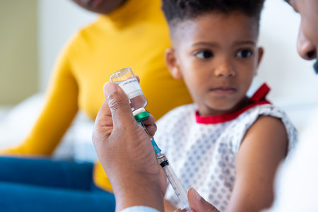 African american male doctor preparing vaccine for boy patient, with mother in hospital. Hospital, medical and healthcare services.の写真素材