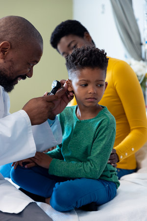 African american male doctor using otoscope to examine ear of boy patient with mother. Hospital, medical and healthcare services.の写真素材