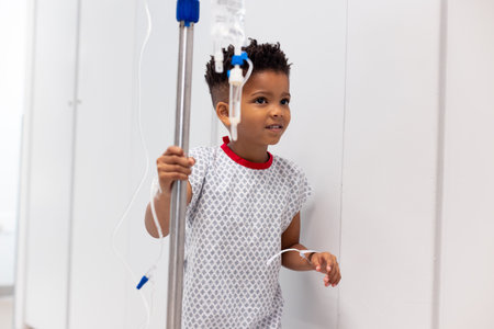 Smiling african american boy patient walking with drip stand in hospital corridor with copy space. Hospital, medical and healthcare services.の写真素材