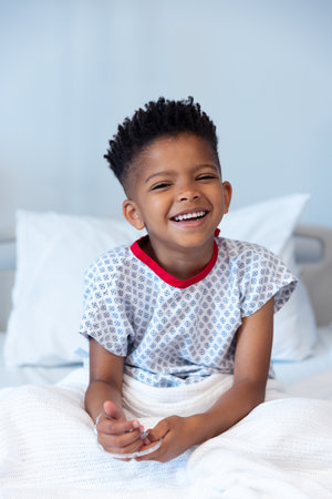 Happy african american boy patient sitting up in hospital bed, smiling with copy space. Hospital, medical and healthcare services.の写真素材