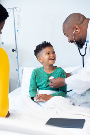 African american male doctor using stethoscope on boy patient in hospital with copy space. Hospital, medical and healthcare services.の写真素材