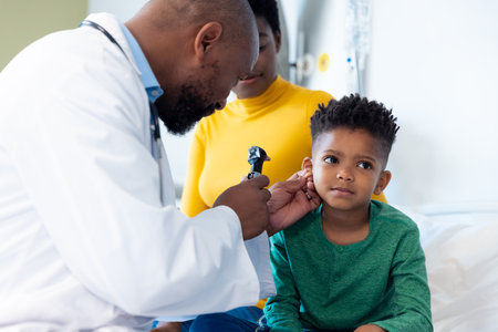 African american male doctor using otoscope to examine ear of boy patient, with mother. Hospital, medical and healthcare services.の写真素材