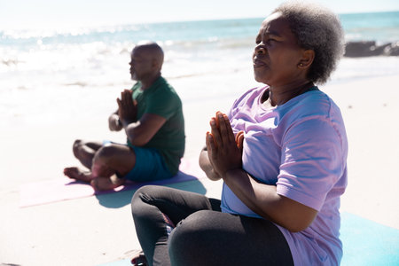 African american senior couple in prayer pose meditating while sitting at beach on sunny day. Unaltered, love, together, yoga, cross legged, retirement, nature, vacation and active lifestyle concept.の写真素材