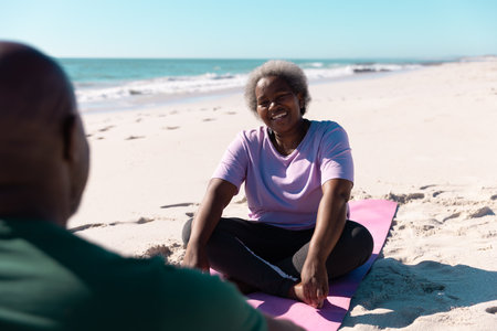 Happy african american senior woman talking with man while relaxing on mats at beach in summer. Copy space, couple, unaltered, love, together, yoga, retirement, nature, vacation and active lifestyle.の写真素材