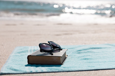 Close-up of book with sunglasses and blue towel on sandy beach against sea during sunny day. Copy space, unaltered, summer, protection, hobby, absence and vacation concept.の写真素材