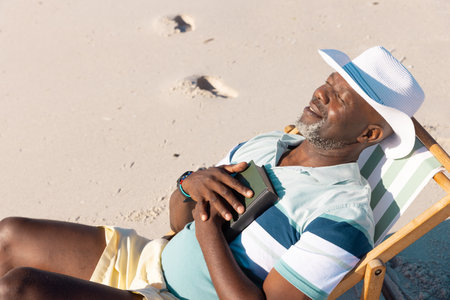 High angle view of african american senior man wearing hat with eyes closed relaxing on deckchair. Beach, summer, book, sunbathing, unaltered, lifestyle, vacation, retirement, enjoyment and nature.の写真素材