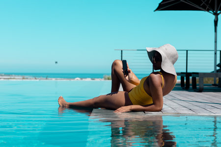 African american wearing straw hat, sitting by swimming pool and using smartphone. Spending quality...