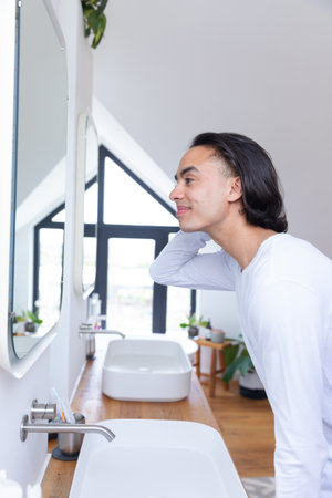 Happy biracial man looking in mirror in bathroom. Spending quality time at home alone, skincare, domestic life and lifestyle concept.の写真素材