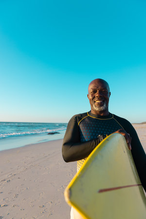 Smiling bald african american senior man carrying surfboard standing at beach against clear blue sky. Copy space, water sports, recreation, retirement, unaltered, vacation, enjoyment, nature, sunset.の写真素材
