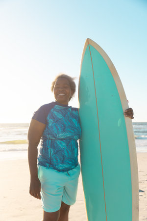 African american senior woman with surfboard standing at beach against sea and sky at sunset. Copy space, water sports, recreation, retirement, unaltered, lifestyle, vacation, enjoyment, nature.の写真素材