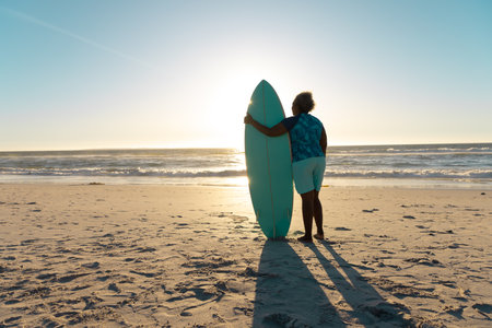 Rear view of african american senior woman with surfboard looking at sea under sky during sunset....
