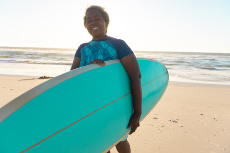Senior african american senior woman with surfboard enjoying on sandy beach against sea and sky. Copy space, water sports, recreation, retirement, unaltered, happy, vacation, sunset, nature.の写真素材