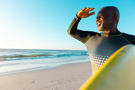 Smiling bald african american senior man shielding eyes and carrying surfboard at beach against sky....