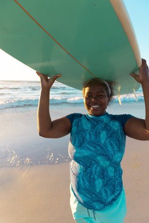 Happy african american senior woman carrying surfboard on head while standing at beach during...