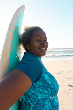 Side view of african american senior woman with surfboard standing at beach against sea and sky. Copy space, water sports, recreation, retirement, unaltered, sunset, vacation, enjoyment, nature.の写真素材