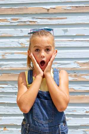 Portrait of surprised biracial girl holding her face with mouth open over blue wall. Spending...