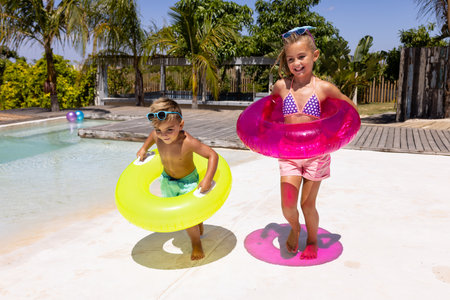Happy biracial brother and sister holding swim rings by the swimming pool. Spending quality time,...
