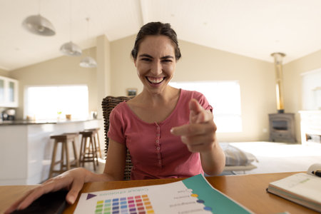 Portrait of cheerful caucasian young woman with color swatch pointing at camera in home office. Copy...
