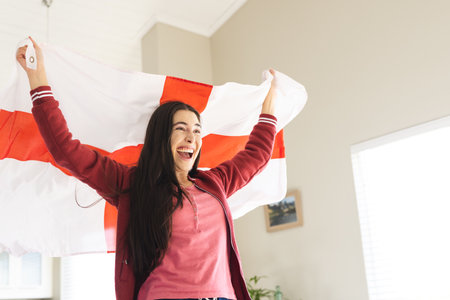 Cheerful caucasian young woman raising flag of england and screaming while watching match at home. Copy space, unaltered, lifestyle, patriotism, fan, sports, support and celebration concept.の写真素材
