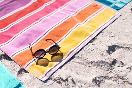 High angle view of sunglasses and colorful towel on sandy beach during sunny day. Copy space, unaltered, protection, vacation, fashion, picnic and summer concept.の写真素材