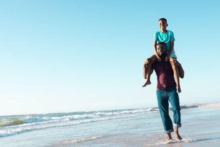 Happy african american father carrying son on shoulders and walking at seashore against clear sky. Copy space, unaltered, family, together, childhood, nature, vacation, enjoyment and summer.の写真素材