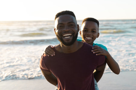Portrait of cheerful african american man piggybacking son against scenic sea and sky at sunset. Copy space, unaltered, family, together, childhood, nature, vacation, enjoyment and summer concept.の写真素材