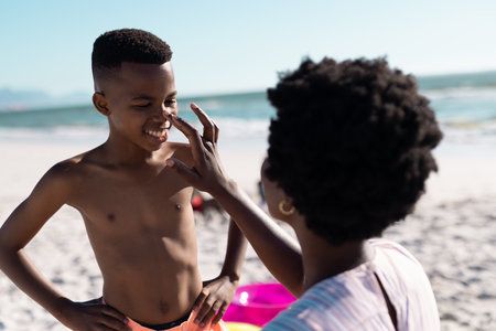 African american mother applying sunscreen on son's nose at beach against clear sky, copy space. Unaltered, family, together, protection, skincare, nature, vacation, enjoyment and summer.の写真素材