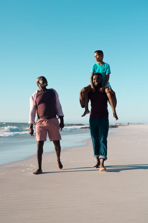 African american man carrying son on shoulders while walking with senior father at beach under sky. Copy space, unaltered, multigeneration family, together, child, nature, vacation, enjoy, summer.の写真素材
