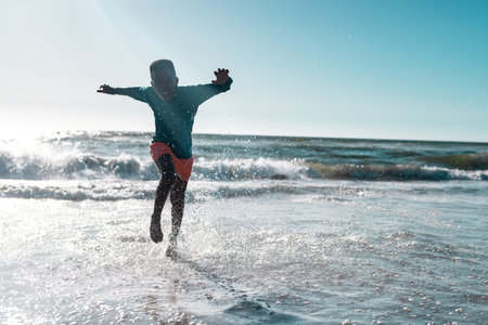 Full length of carefree african american boy running and splashing water in sea against clear sky. Copy space, unaltered, childhood, nature, vacation, playing, enjoyment and summer concept.の写真素材