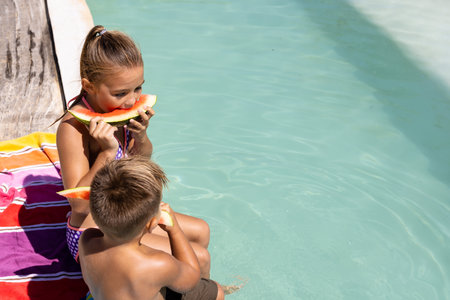 Happy biracial brother and sister eating watermelon by the swimming pool. Spending quality time, lifestyle, family, childhood, summertime and vacation concept.の写真素材
