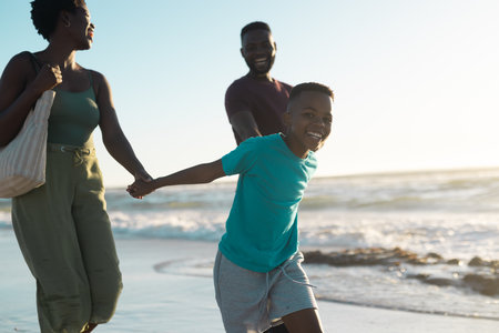 African american cheerful boy holding mother and father's hands and walking in sea against sky. Copy space, unaltered, family, together, childhood, nature, vacation, enjoyment and summer concept.の写真素材