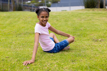 Portrait of happy african american schoolgirl sitting on grass at school. School, education and learning concept.の写真素材