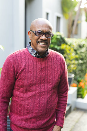 Portrait of happy senior african american man wearing glasses and red jumper standing outside house. Senior lifestyle, summer and nature, unaltered.の写真素材