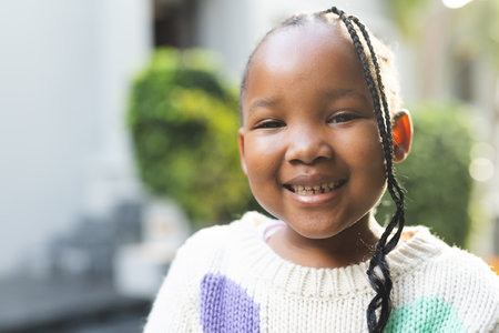 Portrait of happy african american girl with braids standing outside house. Childhood, summer and nature, unaltered.の写真素材