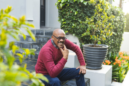 Portrait of happy senior african american man wearing glasses sitting on stairs outside house. Senior lifestyle, summer and nature, unaltered.の写真素材