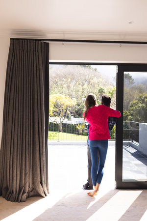Happy diverse couple embracing and looking through window at home, copy space. Lifestyle, togetherness, relationship and dometic life, unaltered.の写真素材