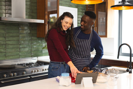 Happy diverse couple baking together in kitchen, using tablet at home. Lifestyle, togetherness, relationship, baking, recipe, communication and domestic life, unaltered.の写真素材