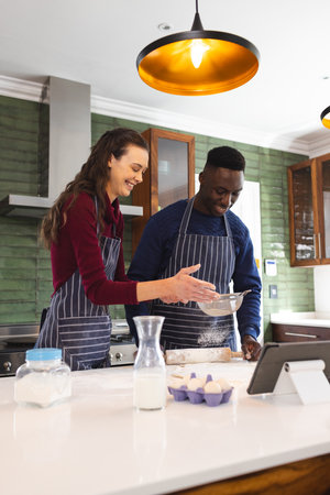 Happy diverse couple baking together in kitchen, using tablet at home. Lifestyle, togetherness,...