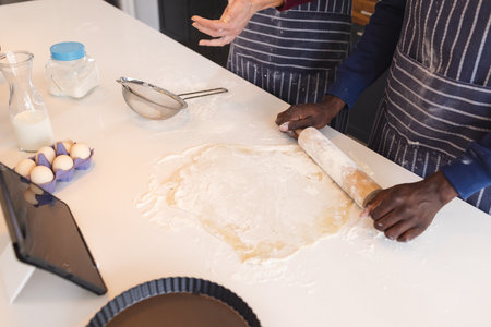 Midsection of diverse couple baking together in kitchen, using tablet at home. Lifestyle, togetherness, relationship, baking, recipe, communication and domestic life.の写真素材