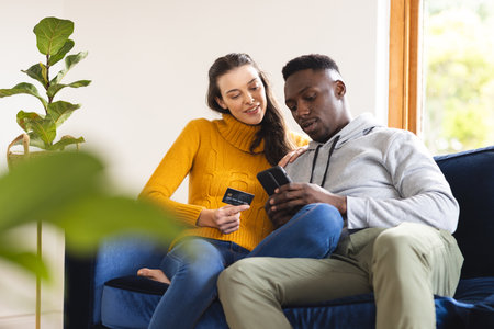 Happy diverse couple sitting on sofa using smartphone for online shopping at home. Lifestyle, togetherness, relationship, online shopping, communication and dometic life, unaltered.の写真素材