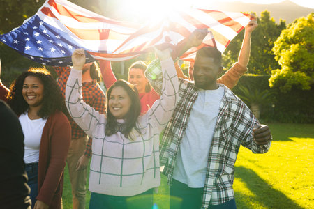Happy diverse male and female friends holding american flag in sunny garden. Celebration, friendship, patriotism, american culture and tradition, unaltered.の写真素材